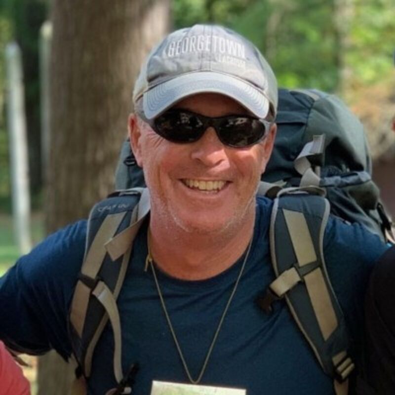 The image shows a man wearing a Georgetown hat, sunglasses, and a backpack. He is smiling and appears to be outdoors, possibly hiking. The background is blurred, suggesting a natural environment. He seems to be enjoying himself.
